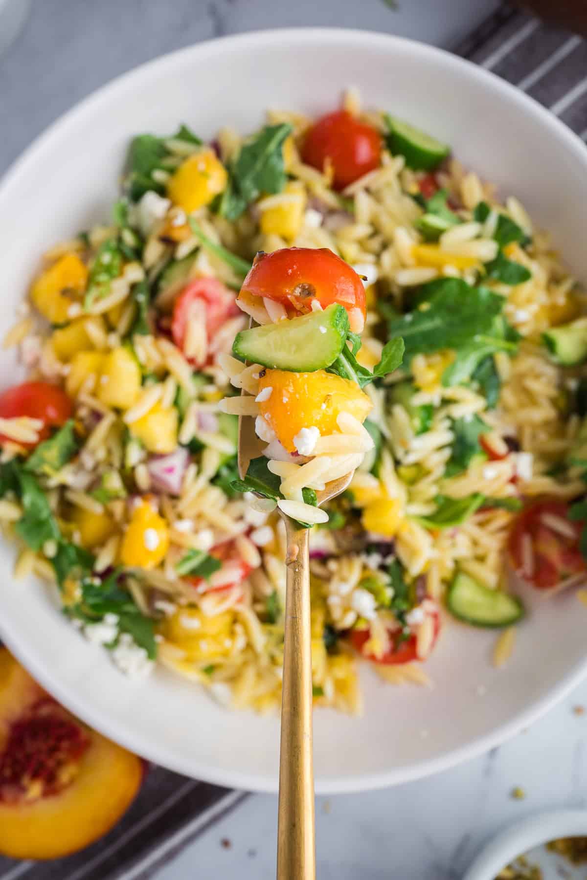 Close-up of a spoon of orzo salad highlighting juicy peaches, creamy feta, cherry tomatoes, cucumbers, and fresh mint.