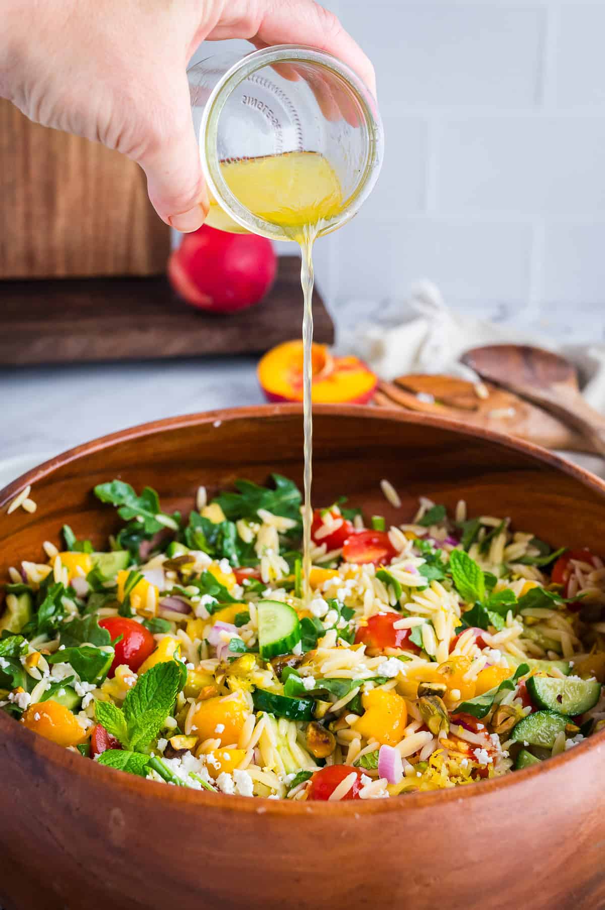 Lemon dressing being poured into summer orzo salad with peaches, tomatoes, mint, and feta.