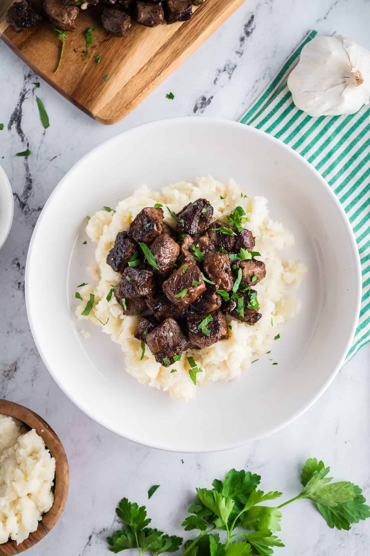 Steak bites over mashed potatoes on a white marble table flanked with a striped kitchen towel. 