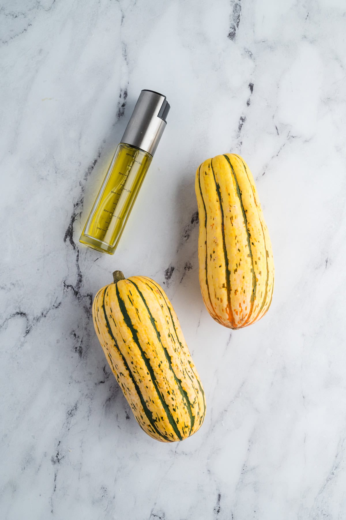 An oil mister and two delicata squash on a marble countertop.