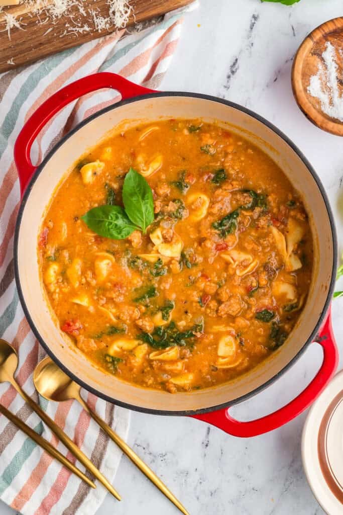 A pot of sausage and tortellini soup on a white marble counter top styled with with gold spoons, a striped napkin, shredded parmesan cheese and salt.