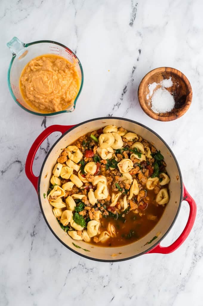 A pot of tortellini soup on a white marble counter with cauliflower puree in a measuring cup.