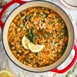 Hearty Tuscan-style white bean soup with kale and vegetables served in a Dutch oven with lemon wedges on top.