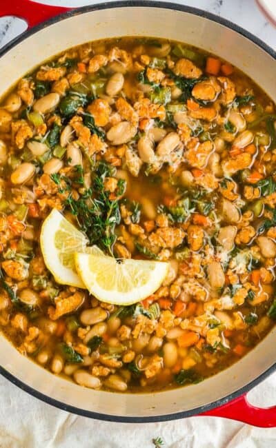 Hearty Tuscan-style white bean soup with kale and vegetables served in a Dutch oven with lemon wedges on top.