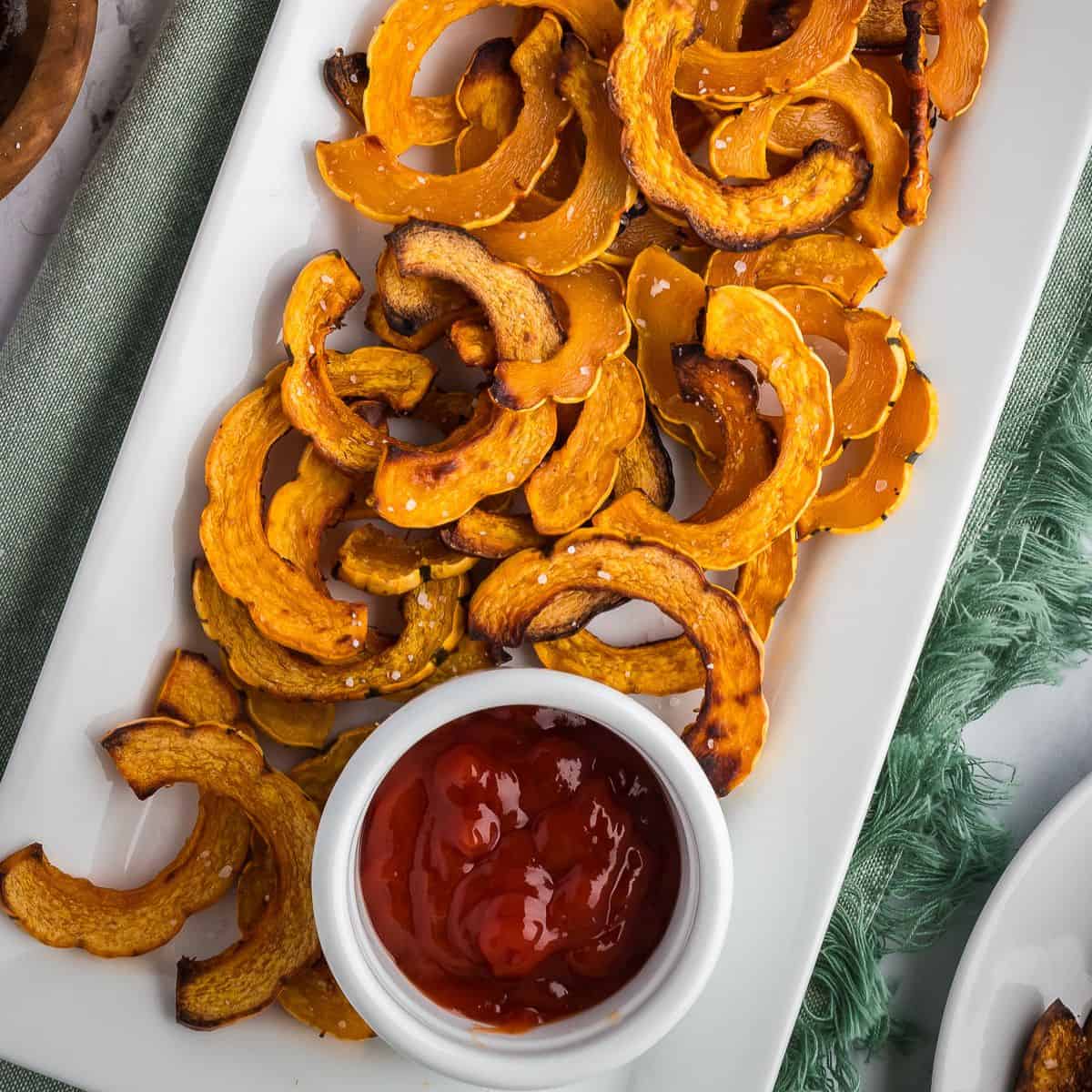 Crispy roasted delicata squash on a white platter with a small ramekin of ketchup.