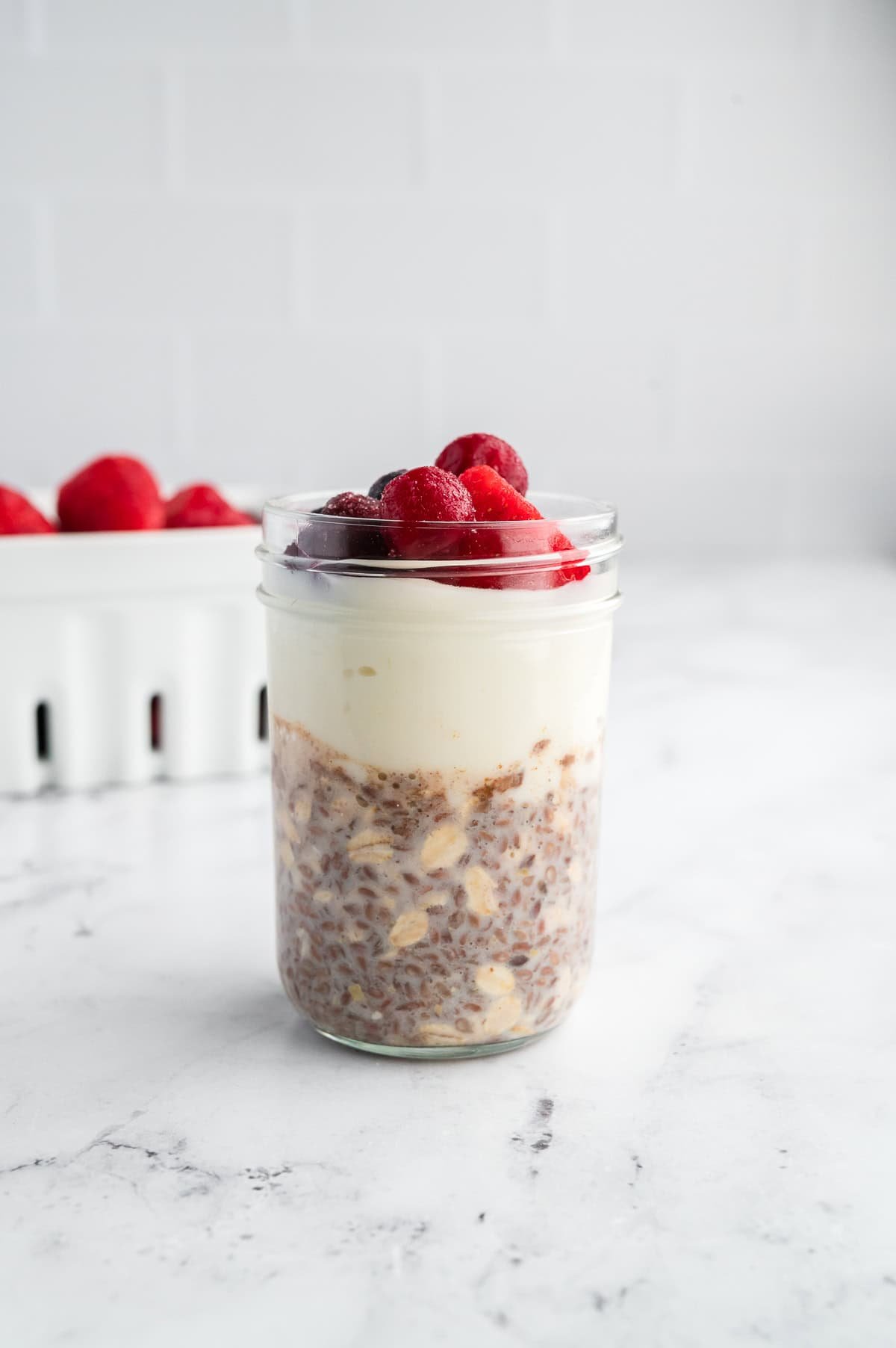 Jar of high-protein overnight oats topped with frozen mixed berries, with a white berry container blurred in the background.