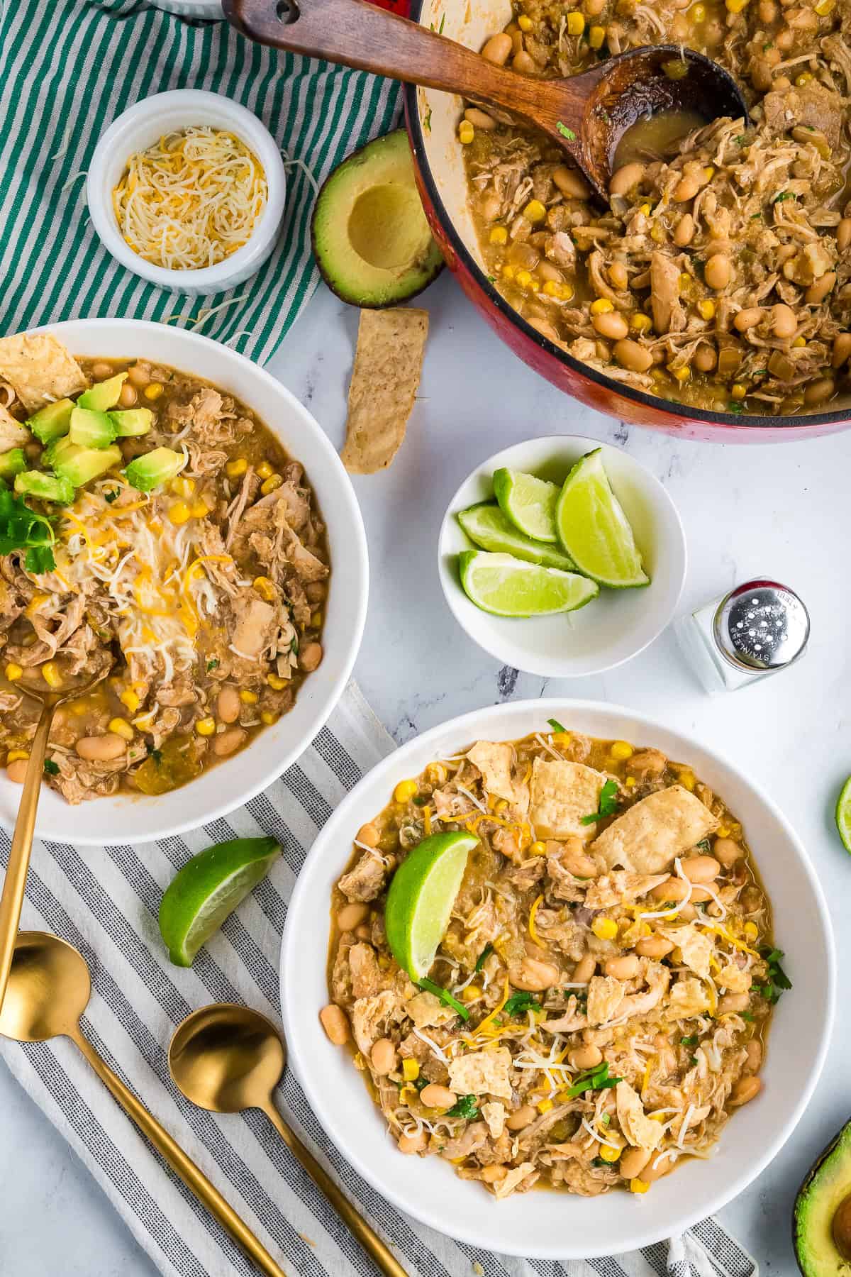 White chicken chili in a red Dutch oven with a wooden ladle, surrounded by avocado, cheese, and lime wedges.