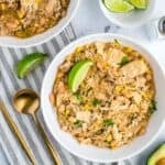 Two bowls of white chicken chili garnished with lime wedges, tortilla chips, cheese, and avocado on a light surface.