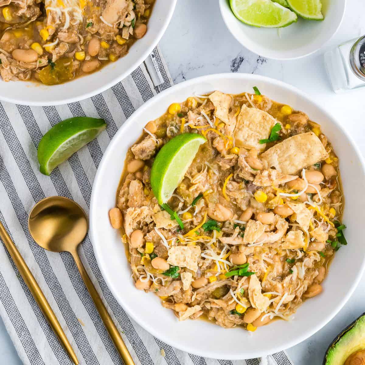 Two bowls of white chicken chili garnished with lime wedges, tortilla chips, cheese, and avocado on a light surface.